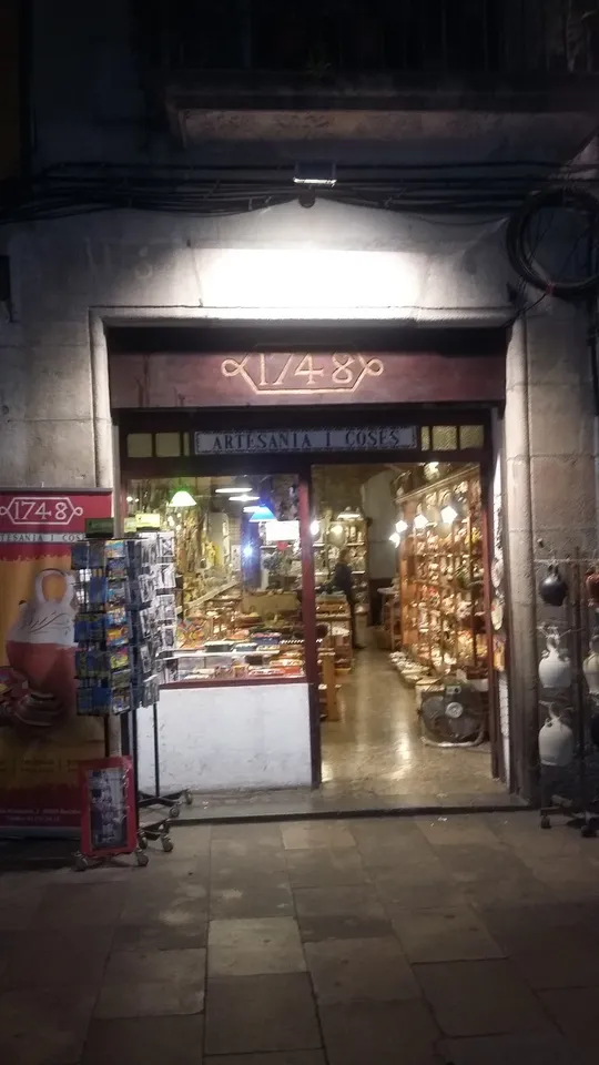 La façana de la botiga de nit, amb el rètol 1748 i Artesania i Coses i els prestatges de ceràmica il·luminats a dins